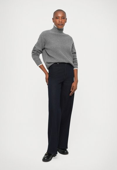 Pull à col roulé gris à côtes, associé à un pantalon large navy taille haute. Le mannequin porte des chaussures noires, les mains sur les hanches. Fond minimaliste.