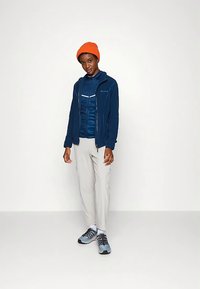 Μπλε μπουφάν με φερμουάρ, μπλε μονωτική στρώση, ανοιχτό γκρι παντελόνι και γκρι-μπλε αθλητικά παπούτσια. Πορτοκαλί πλεκτός σκούφος. Απλή, λειτουργική ενδυμασία για υπαίθριες δραστηριότητες.