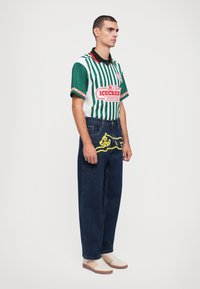Green and white striped short-sleeve jersey with a black collar, denim jeans featuring a yellow graphic, and cream sneakers with brown accents.