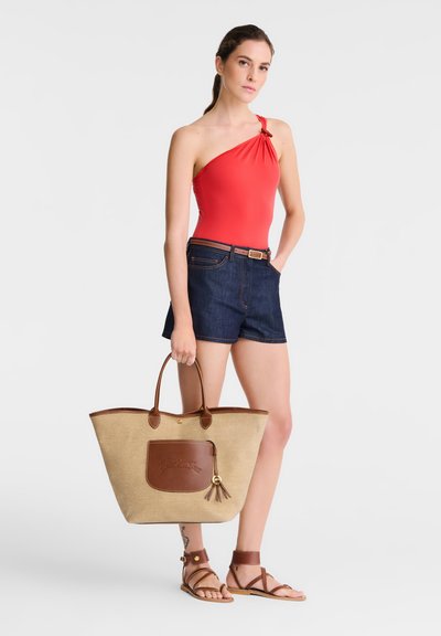 Red one-shoulder swimsuit, dark denim shorts, brown belt, tan straw tote bag with leather accents and tassel, brown strappy sandals.
