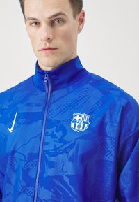 Blauwe sportjacket met hoge kraag, gladde textuur, ritssluiting, met een wit Nike-logo en het FC Barcelona-embleem op de borst.