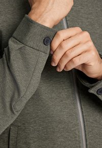 Chaqueta verde oliva que destaca por su diseño ajustado, tejido texturizado y puños abotonados. Presenta una cremallera visible y herrajes contrastantes.