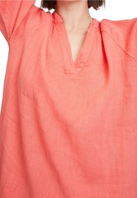 Coral linen tunika dengan leher V dan detail kerut di leher; potongan santai dengan lengan panjang yang sedikit menggelembung.