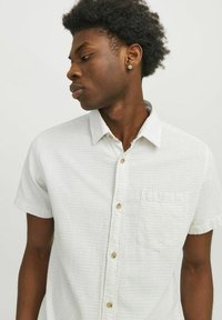 Camisa de manga corta abotonada en tejido ligero, con un sutil patrón de puntos, botones beige y un bolsillo en el pecho. El color es predominantemente blanco.