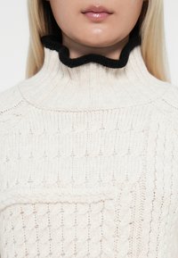 Krämfärgad stickad turtleneck-tröja med ribbat mönster och en svart volangkrage. Har ett flätat mönster genom hela kroppen.