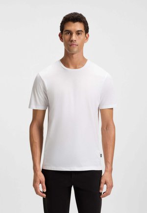 Mann steht frontal, trägt ein einfarbiges weißes T-Shirt mit kurzen Ärmeln und schwarze Hosen vor einem einfarbigen weißen Hintergrund.