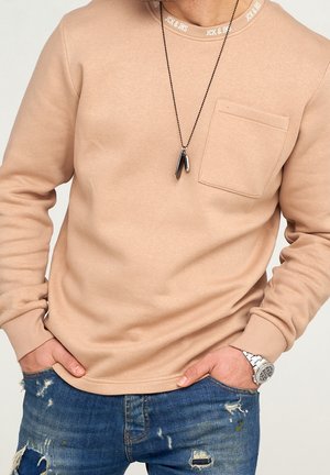 Sweatshirt beige avec un col ras du cou côtelé, doté d'une poche poitrine et de manches longues, associé à un jean bleu déchiré. Collier en argent visible.