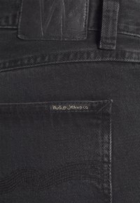 Svarta denimjeans med en strukturerad midja, en läderpatchdetalj och ett subtilt vågformat broderimönster på fickan.