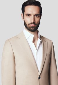 Beżowa lniana marynarka o usztywnionej konstrukcji, z klapami, zapięciem na jeden guzik i kieszenią naszywaną, noszona na białej koszuli z kołnierzykiem.