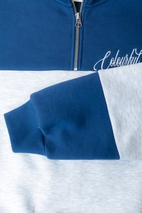 Blauwe en grijze zip-up sweatshirt met ribgebreide manchetten. Heeft een bovenste blauwe paneel, een onderste grijze paneel, en geborduurde tekst op de voorkant.