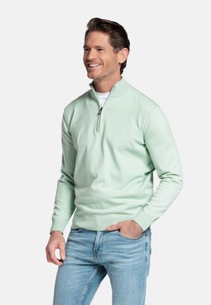 Lachende man met kort bruin haar, gekleed in een licht muntgroene trui met halve rits over een wit overhemd en lichtblauwe spijkerbroek, met de hand in de zak.