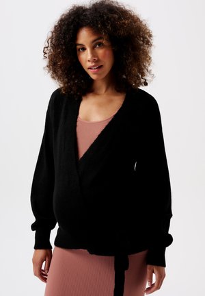 Zwarte wikkelcardigan met lange, opbollende mouwen en een strikdetail in de taille, gedragen over een geribde roze tanktop. Zachte textuur, losse pasvorm.