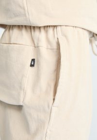Beige kordfloretbyxor med elastisk midja, med sidofickor och en liten svart etikett på framsidan nära fickans söm.