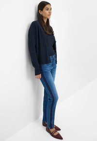 Donna appoggiata a una parete bianca, indossa un cardigan blu navy, una maglietta a coste, jeans blu con una striscia laterale scura e ballerine porpora a punta con fibbie.