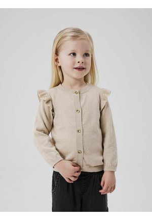 Cardigan bej tricotat cu accente volan pe umeri, nasturi aurii și manșete cu fâsle. Copilul îl poartă cu pantaloni întunecați, zâmbind.