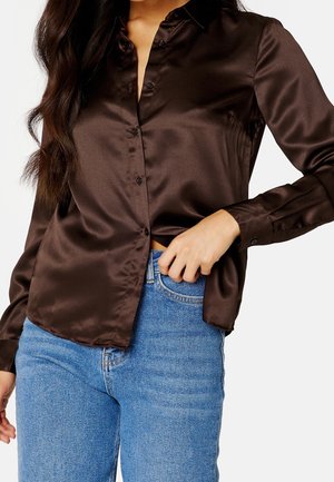 Femme portant une chemise en satin brun foncé boutonnée, partiellement rentrée dans un jean bleu clair, ajustant la chemise à la taille d'une main.