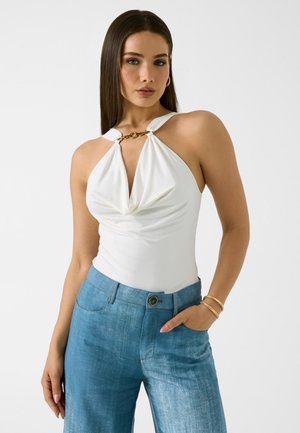 Femme aux cheveux raides et foncés portant un haut halter blanc avec un détail de chaîne dorée et un jean taille haute bleu, debout avec une main dans la poche.