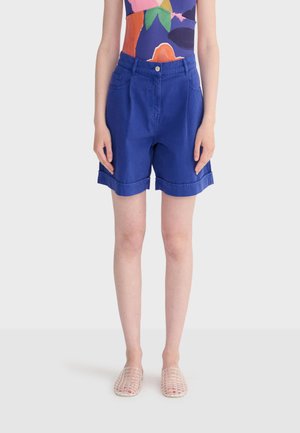 Persoon die felblauwe opgerolde korte broek, kleurrijk patroonloze mouwloze top en transparante geweven instappers draagt, staand met ontspannen armen.