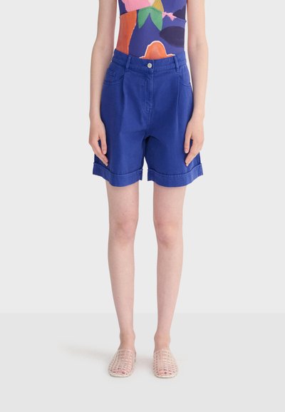 Personne portant un short bleu vif retroussé, un haut sans manches à motifs colorés, et des chaussures transparentes tissées sans lacets, debout les bras détendus.