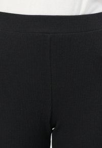 Gros plan sur la taille et la partie supérieure de la cuisse d’un pantalon en tissu noir côtelé, porté avec un top blanc côtelé rentré.