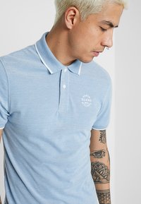 Lyseblå polo-skjorte med teksturert mønster, hvite krageakenter og en liten logo på venstre bryst. Kortermet og fitted design.