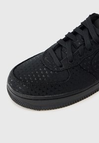 Μαύρο αθλητικό παπούτσι με υφασμάτινη επιφάνεια που διαθέτει μικρά διάτρητα σχέδια, στρογγυλή μύτη, επίπεδα κορδόνια και ανθεκτική σόλα από καουτσούκ.