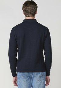 Tengerészeti hosszúujjú póló, textúrált kötött mintával. Kerek nyakkal és bordázott mandzsettákkal rendelkezik, világoskék farmernadrággal párosítva.