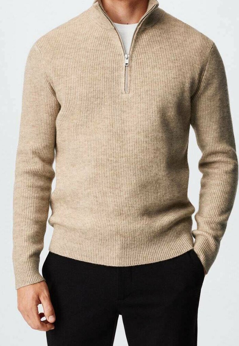 Uomo che indossa un maglione beige a coste con zip a quarti e pantaloni neri, con una mano in tasca, su uno sfondo neutro.