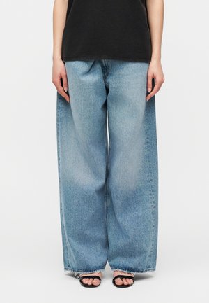 Person trägt weite, hellblaue Jeans aus Denim, ein schwarzes ärmelloses Oberteil und schwarze offene Sandalen vor einem schlichten weißen Hintergrund.