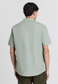 Mann, der sich abwendet und ein hellgrünes Kurzarmhemd sowie eine dunkelgrüne Hose trägt, vor einem schlichten hellen Hintergrund.