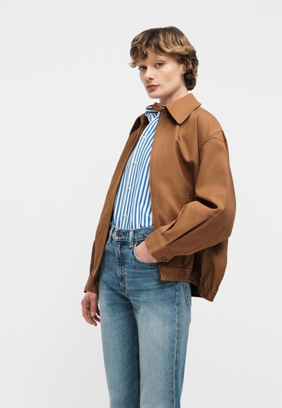 Άτομο που φοράει ένα καφέ τζάκετ με φερμουάρ πάνω από ένα μπλε και άσπρο ριγέ πουκάμισο με κουμπιά, μαζί με ανοιχτόχρωμα τζιν, στέκεται με το ένα χέρι στην τσέπη.