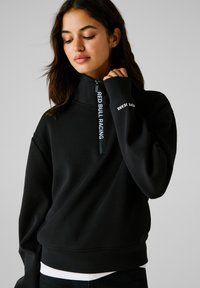 Mlada ženska v črni majici z zadrgo, na zadrgi napis "RED BULL RACING" in na rokavu napis "PEPE JEANS", zamišljeno gleda navzdol.