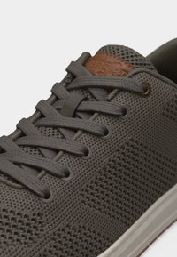 Κοντινό πλάνο γκρι πλεκτού αθλητικού παπουτσιού με κορδόνια ίδιου χρώματος, καφέ δερμάτινη ετικέτα στη γλώσσα και σχεδιασμό με διάτρητο ύφασμα.