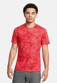 Camiseta deportiva roja con un patrón texturizado, mangas cortas y cuello redondo. Presenta un pequeño logo dorado en la parte superior izquierda.
