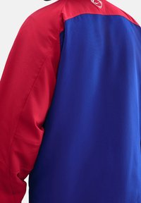 Roodblauwe jas met een kleurblokdesign, gladde textuur en een logo op de achterkant, zichtbare stiksels op de mouwen.