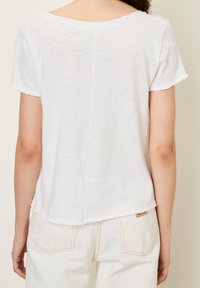 Kvinna som bär en vit kortärmad skjorta och vita byxor, sedd bakifrån mot en enfärgad ljus bakgrund.