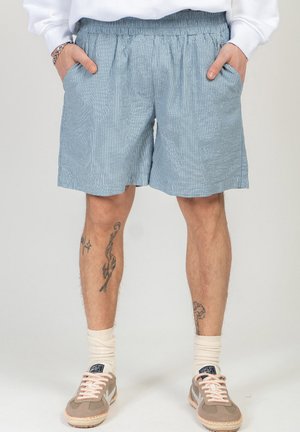 Mężczyzna w jasnoniebieskich szortach w paski, białych skarpetach do kostek i beżowych trampkach, z rękami w kieszeniach i widocznymi tatuażami na nogach.