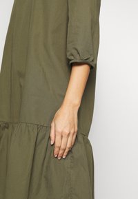 Abito verde oliva con orlo arricciato e svasato e maniche a tre quarti. Tessuto morbido e liscio con una sottile trama. Mano appoggiata sulla cucitura laterale.