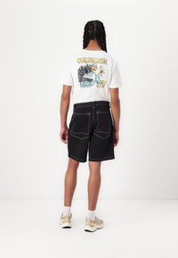 Sorte denimshorts med hvite sømmer, kombinert med en hvit t-skjorte med grafikk og flerfarget trykk på baksiden. Beigesneakers med hvite sokker.