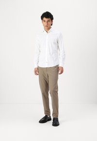 Cămașă albă cu mânecă lungă, cu un model subtil, asortată cu pantaloni bej și pantofi negri. Cămașa are un design cu nasturi.