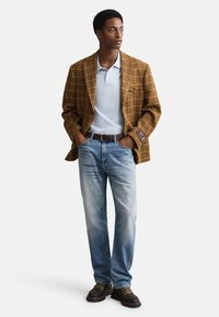 Bruine geruite blazer over een lichtblauw poloshirt, gecombineerd met vervaagde blauwe jeans en zwarte wandelschoenen. De outfit heeft een ontspannen pasvorm.