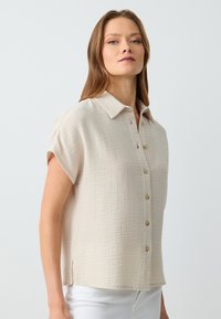 Donna che indossa una camicia beige a maniche corte con texture e bottoni, abbinata a pantaloni bianchi, in piedi contro uno sfondo chiaro e semplice.