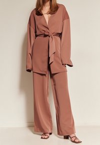 Woman wearing mauve wide-leg pants, matching belted wrap jacket, and brown block-heeled sandals against plain background.