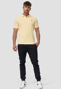Gul polo-skjorte med krage og korte ermer, kombinert med svarte smale bukser og hvite joggesko med svart detaljer.