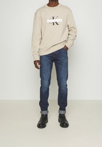 Sudadera beige con logo "Calvin Klein Jeans", jeans azul oscuro y botines negros, mostrados en un modelo sobre un fondo neutro.