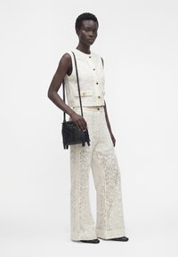 Femme portant un gilet sans manches en dentelle blanche et un pantalon large, tenant un sac bandoulière noir à franges, debout devant un fond blanc uni.