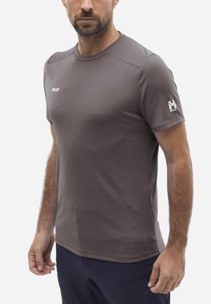 Homme portant un t-shirt gris texturé à manches courtes ajusté de la marque Millet et un pantalon sombre, debout les bras détendus le long du corps.