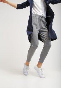 Graue Jogginghose mit elastischem Bund und Kordelzug. Kombiniert mit einem weißen Hemd und einem langen, marineblauen Mantel. Weiße Sneakers runden das Outfit ab.