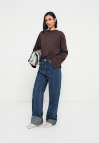 Chemise marron oversize à manches longues, jean bleu taille haute à jambes larges, chaussures beige à enfiler, et une pochette argentée à motif serpent.