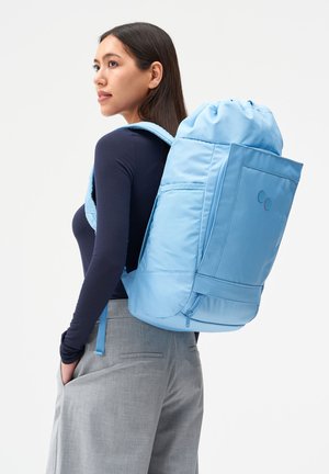 Frau trägt ein marineblaues Langarmshirt und graue Hose, hat einen großen hellblauen Rucksack über der Schulter und schaut nach links.